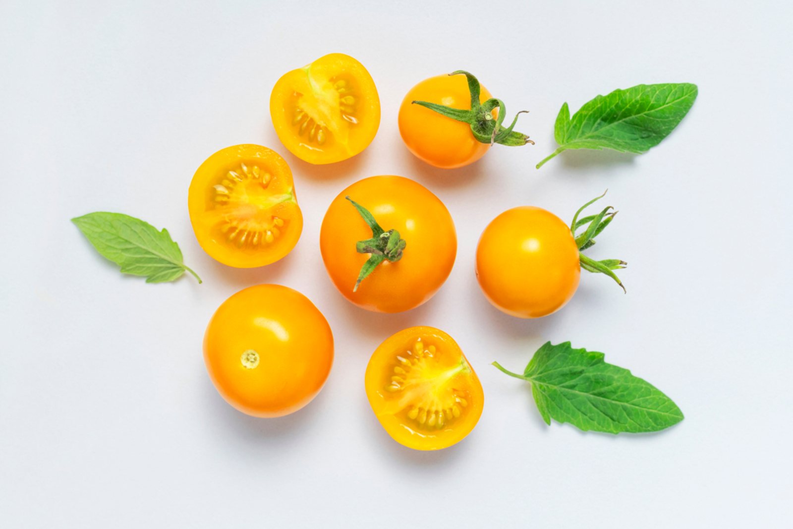Whole and halved yellow-orange tomatoes with green leaves on white