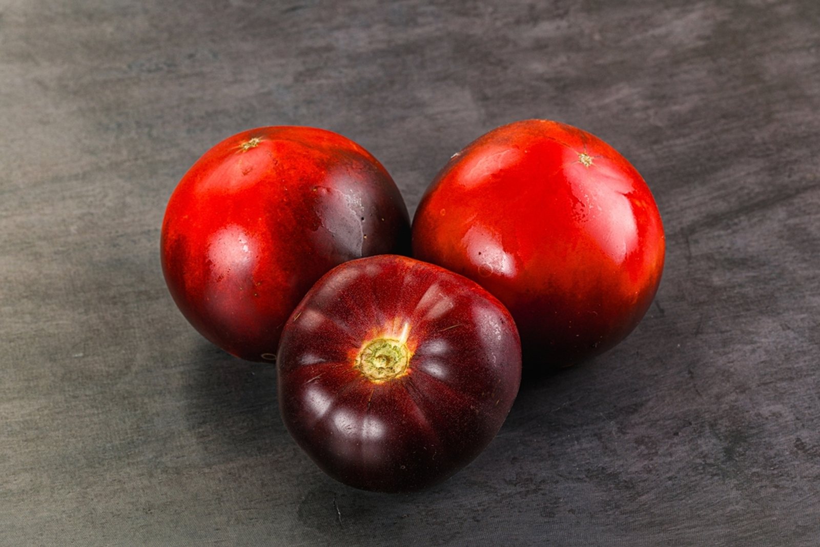 Three dark red-purple tomatoes at full color on a dark surface
