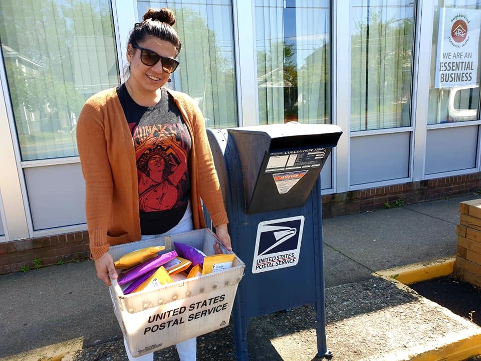 A person carrying a postal crate filled with colorful AONSK orders beside a United States Postal Service collection box