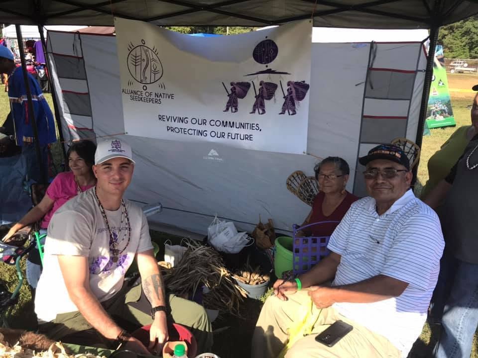 Frank Cain seated with community members beneath an Alliance of Native Seedkeepers tent